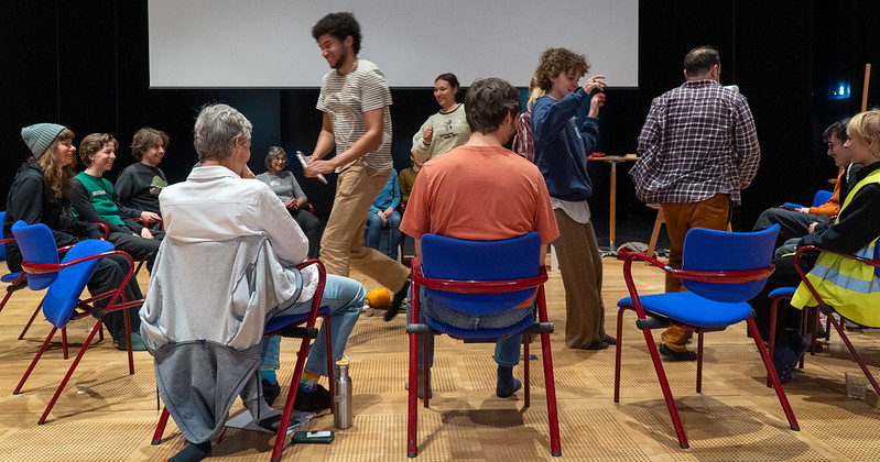 Eine Gruppe von Menschen sitzt aus Sessel im Kreis 2 Personen stehen in der Mitte