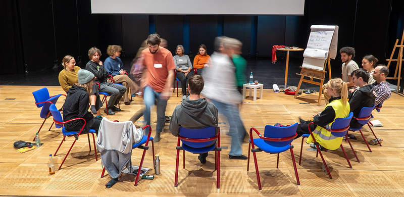 Eine Gruppe von Menschen sitzt aus Sessel im Kreis 2 Personen stehen in der Mitte